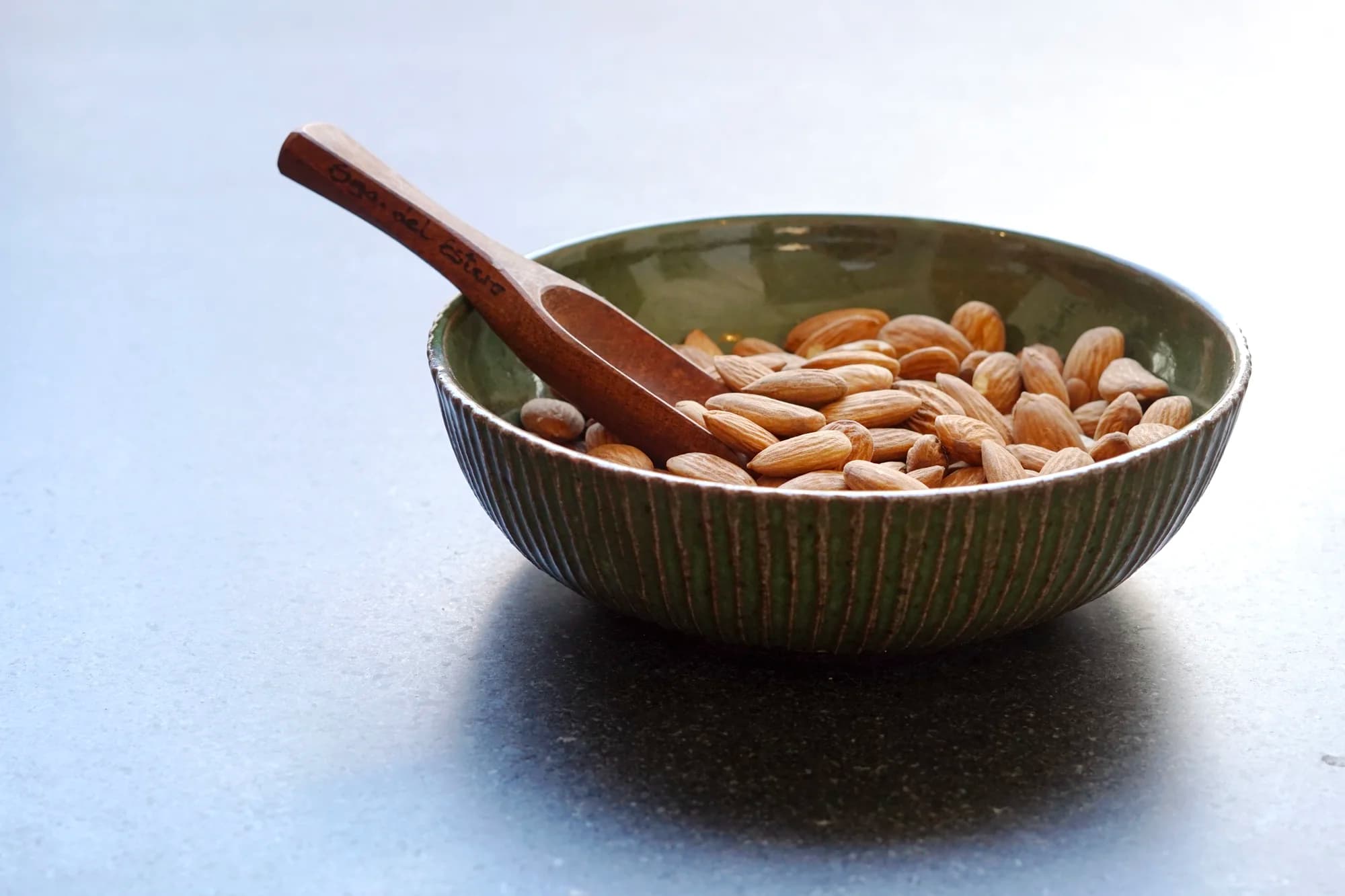 A handmade green ceramic bowl filled with raw almonds and a small wooden scoop