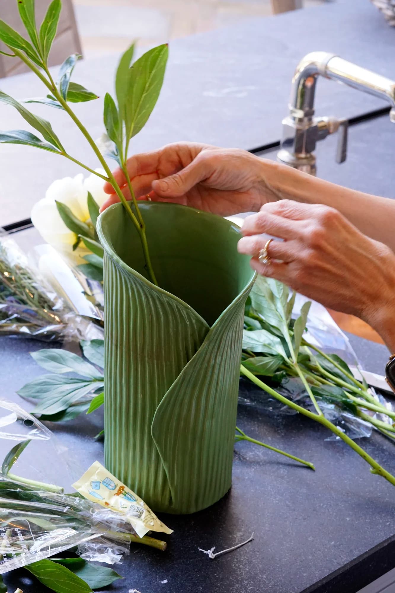 Hands arranging leafy green stems into a tall, ridged green ceramic vase