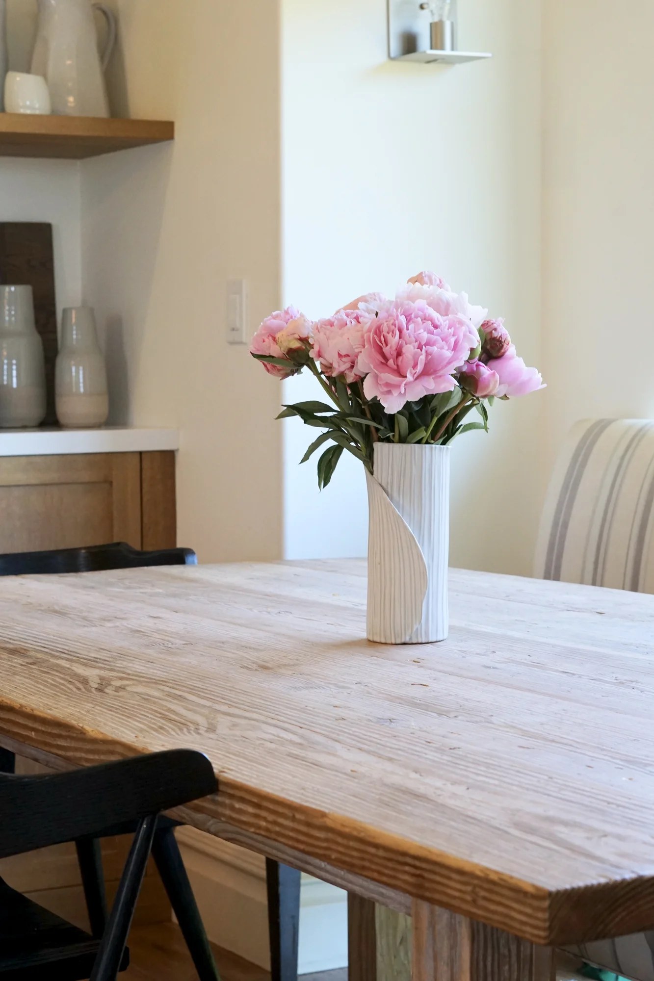 A stoneware vase holding soft pink peonies atop a rustic wooden dining table