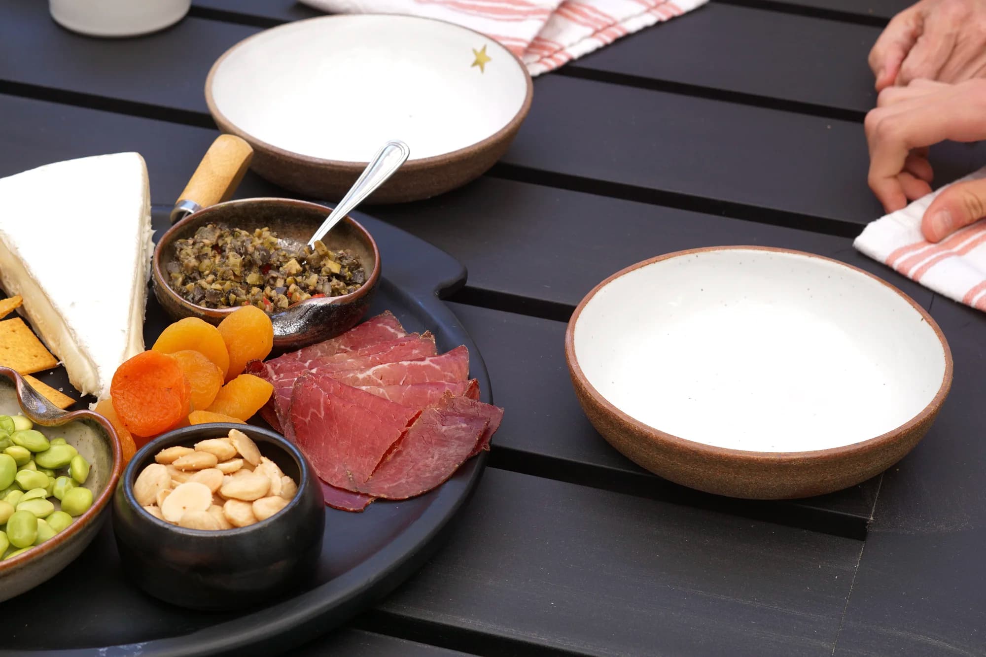 A dark serving platter arranged with cheese, dried apricots, cured meats, nuts, and tapenade