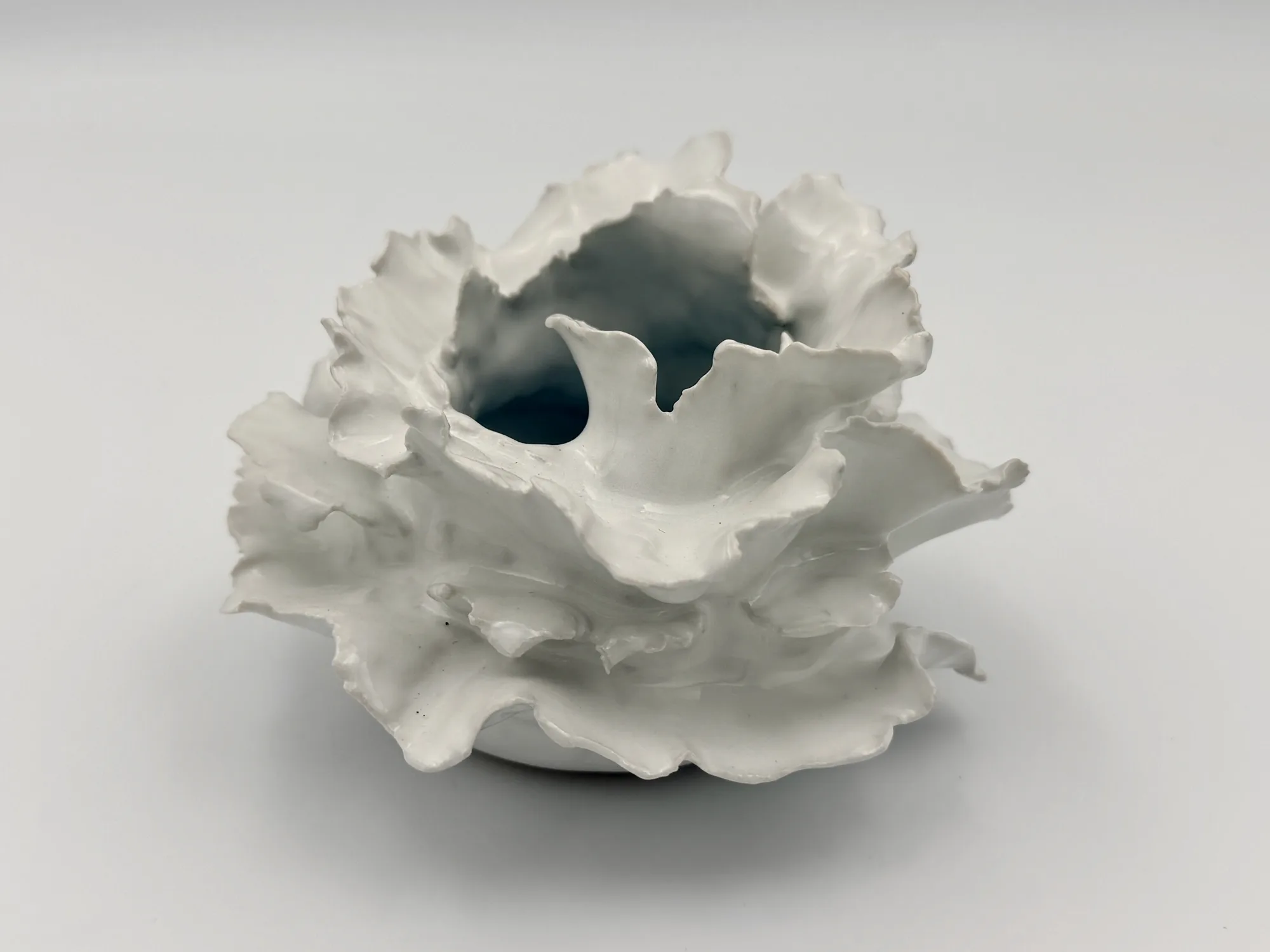 A white ruffled porcelain sculpture with an open, wave-like rim on a soft grey surface